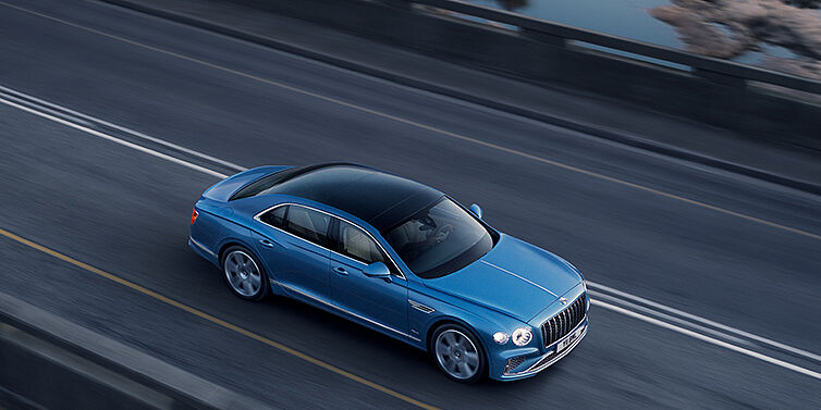 Bentley Oslo New Bentley Flying Spur Azure sedan in Light windsor blue paint by Mulliner, with 22 inch Azure wheels seen from overhead driving dynamically on a country road